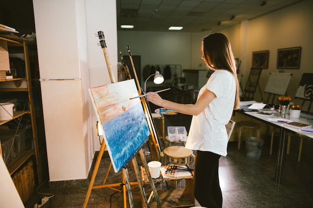 Woman artist painting a picture on easel with oil paints in her workshop