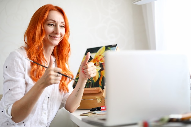 Photo the woman artist sits at the table and shows ok gesture in the laptop screen the artist