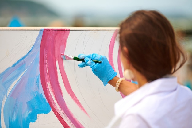 Woman artist working on abstract oil painting with pink and blue colors, hand moving paint brush. Girl creating modern masterpiece. Canvas standing in open-air gallery, creative workshop outdoor