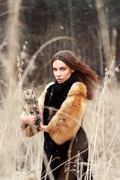 Woman in autumn in fur coat with owl on hand first snow. Beautiful brunette girl with long hair in nature, holding an owl. Romantic, delicate look girls