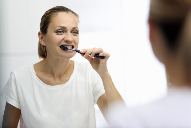 Photo woman in awhite t-shirt is brushing her teeth in front of mirror