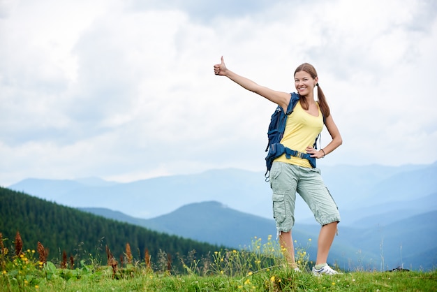 Woman backpacker hiking mountain trail, walking on grassy hill, wearing backpack, using trekking sticks. Hiker enjoying summer day