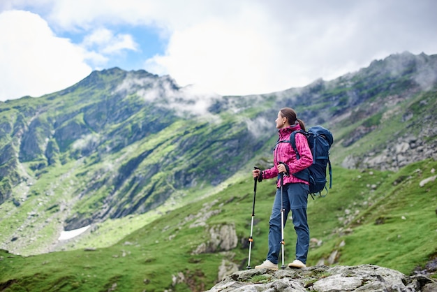 Viaggiatore con zaino e sacco a pelo della donna che riposa mentre facendo un'escursione in piedi sopra una roccia che gode del paesaggio fantastico della montagna intorno