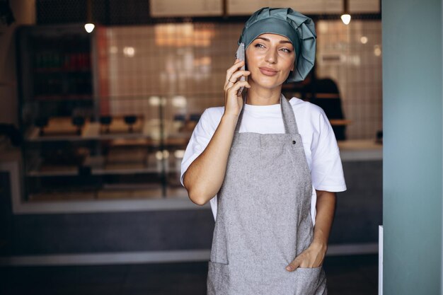 Woman baker in apron standing with phone by the doors at the coffee shop