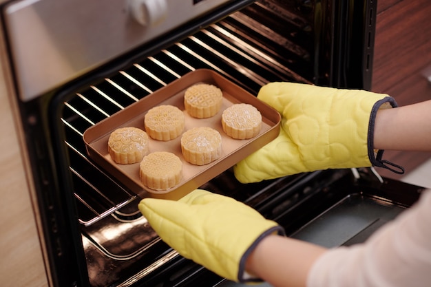 Woman in baking gloves putting tray with mooncakes in preheated oven when cooking delicious dessert ...