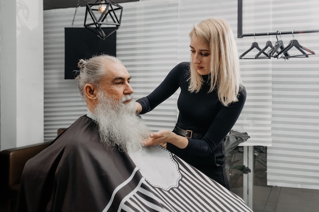 Woman barber cutting hair to an aged bearded man