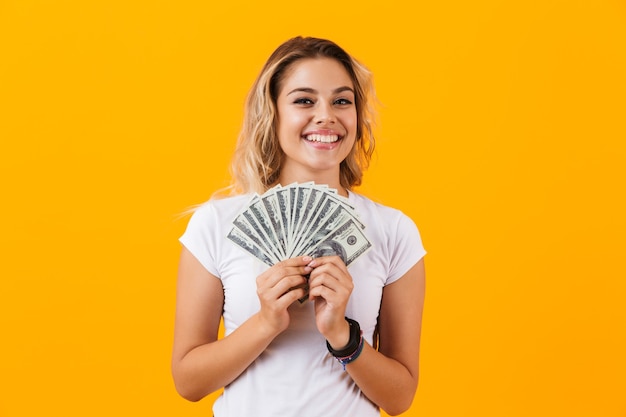 Woman in basic clothing holding fan of dollar money, isolated over yellow wall