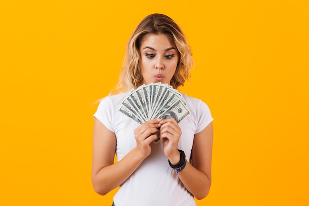 Woman in basic clothing holding fan of dollar money, isolated over yellow wall