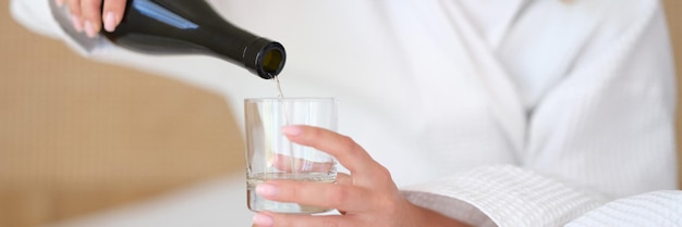 Woman in bathrobe on bed pours water or white wine into glass alone