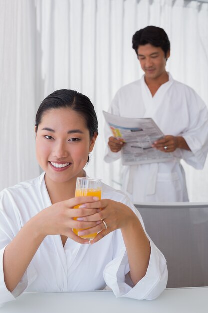 Woman in bathrobe having orange juice with boyfriend in background