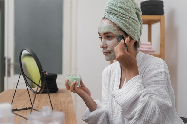 woman in bathrobe skincare with mask applied using a brush reflection in the mirror