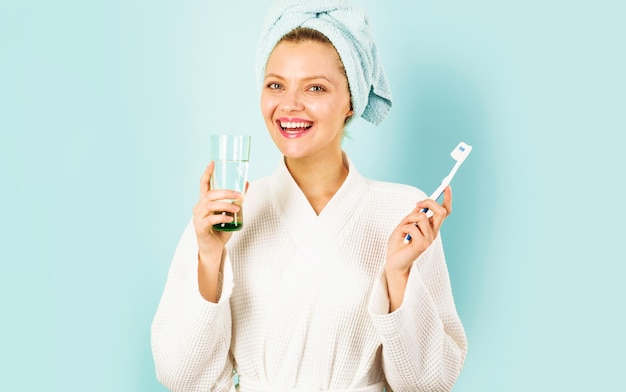 Woman in bathrobe and towel on head brushing teeth holds glass water morning routine dental care