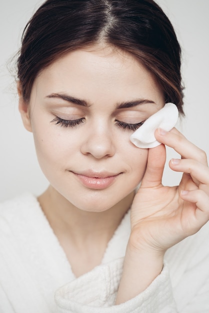 Woman in a bathrobe wipes her face with a soft sponge clean\
skin cosmetology