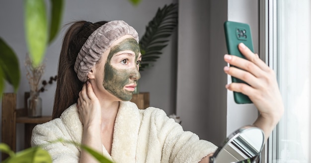 Woman in a bathrobe and with a green cosmetic mask on her face is taking a selfie on her mobile phone