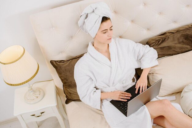 A woman in a bathrobe and with a towel on her head after a shower on the bed with a laptop working and learning. Online work at home. Stay at home during quarantine