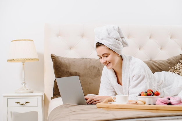 Женщина в халате и с полотенцем на голове после душа на кровати с ноутбуком работает и учится. Онлайн работа на дому. Оставайтесь дома во время карантина