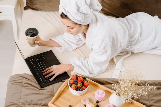 A woman in a bathrobe and with a towel on her head after a shower on the bed with a laptop working and learning. Online work at home. Stay at home during quarantine