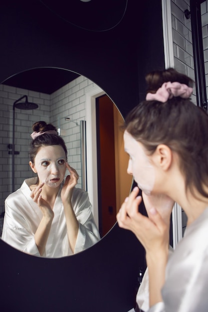 Woman in the bathroom in front of the mirror puts on a rejuvenating face mask from wrinkles