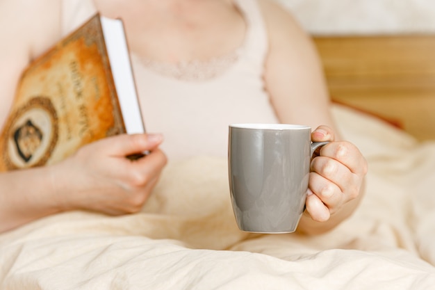 ティーカップと本が付いているベッドの女性。大人の女性がベッドで読書。