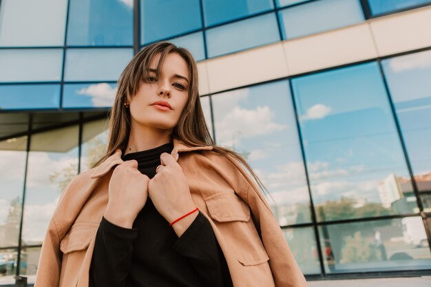 A woman in a beige jacket stands in front of a glass building