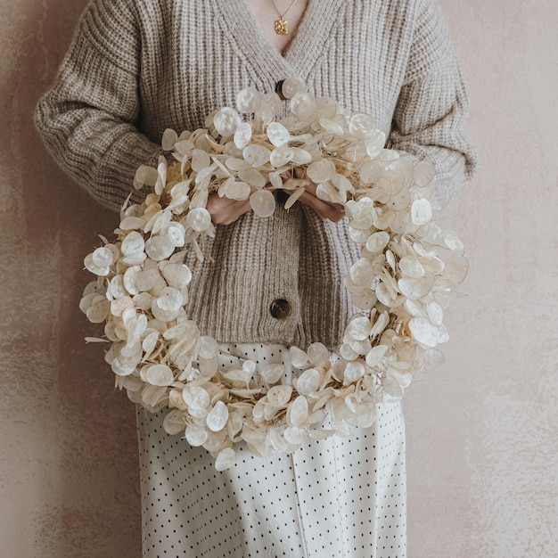 Woman in beige sweater and skirt hold christmas wreath of gold
lunaria leaves in front of neutral dusty pink wall elegant
aesthetic minimalist fashion background