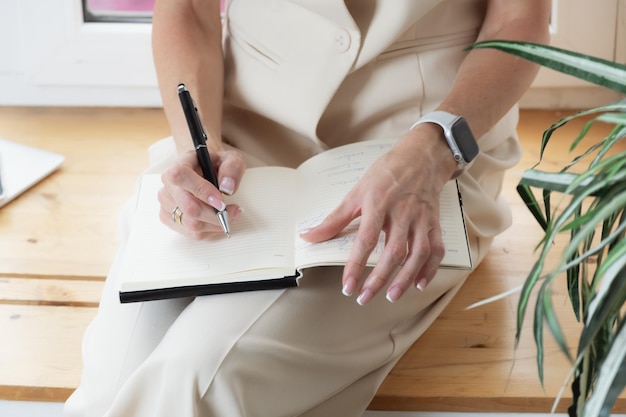 Woman in a beige trouser suit sits on the windowsill in her office and takes notes in a notebook Business woman plans her schedule in a diary