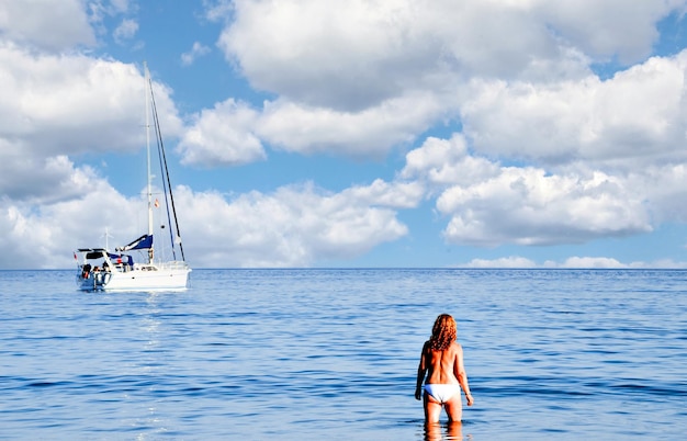 Woman in bikini on the beach looking at the horizon on a blue sky day with white clouds.