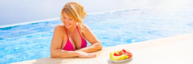 Woman in bikini standing at poolside next to plate of fresh fruit