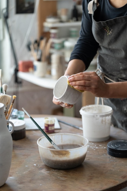 Woman in black apron creates white ceramic mug standing at round wooden table on blurred background