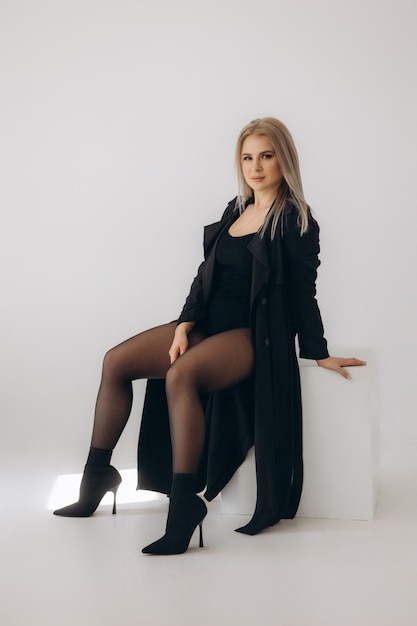 A woman in a black coat and black boots sits on a white chair.