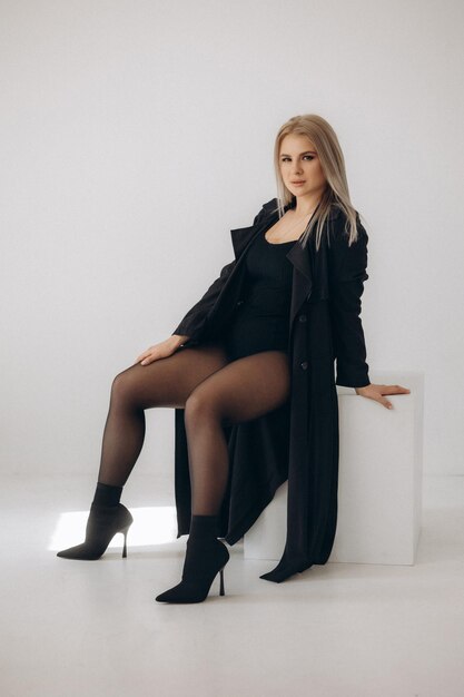 A woman in a black coat and black tights sits on a white cube.