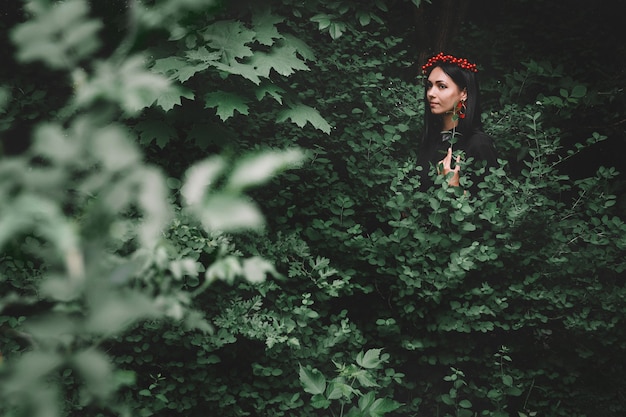 Woman in black dress and red decorations that holds a twig in her hand against background of forest