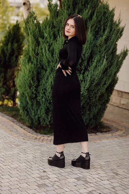 A woman in a black dress stands in front of a tree