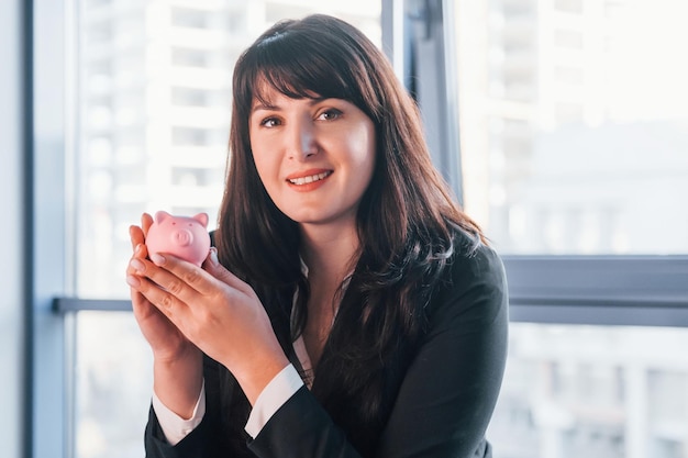 Woman in black formal clothes is indoors in the modern office holds pink piggy bank