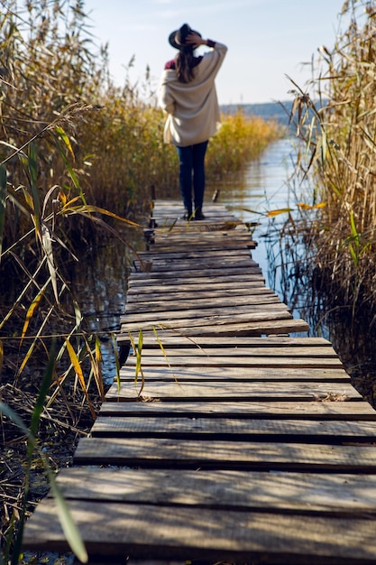 Photo woman in a black hat and sweater stands on the old pier to the bushes by the lake