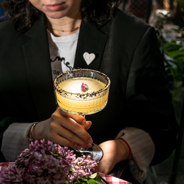 Woman in black is holding cold yellow cocktail which decorated piece of dried flower