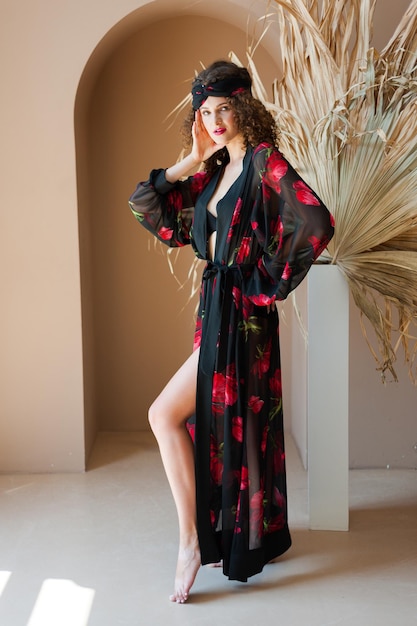 A woman in a black kimono with red roses on it stands in front of a white vase with a feather in the background.