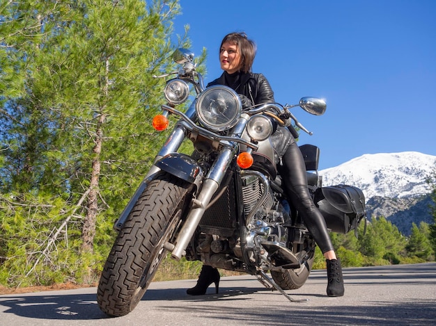 A woman in a black leather biker jacket on a chopper motorcycle in Greece on a road in the forest
