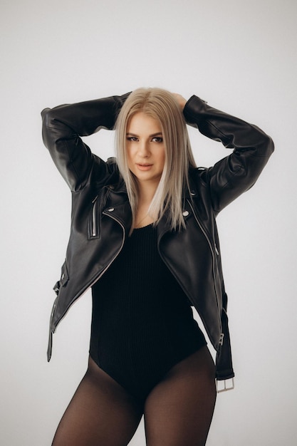 A woman in a black leather jacket with a black shirt and black pants poses with her hands behind her head.