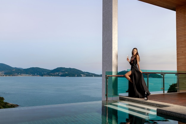 Foto donna in un lungo abito nero si erge con un bicchiere di champagne vicino alla piscina a sfioro