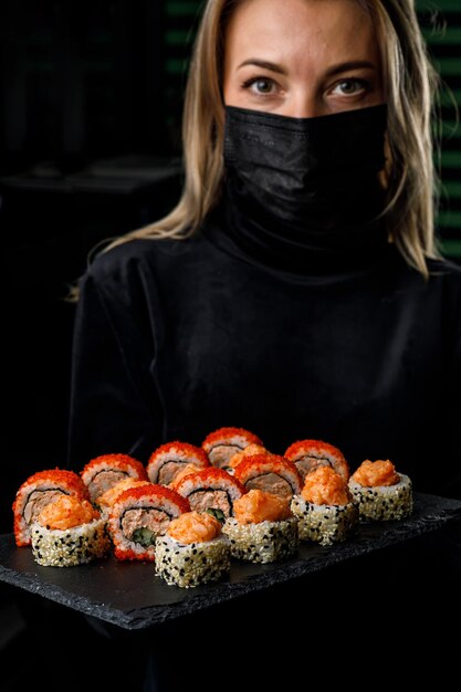 A woman in a black mask holds a tray of sushi.