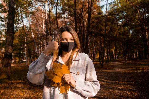 Woman in black medical mask thumbs up and holds bouquet of orange maple leaves. Autumn park or forest on background. Fall season concept. Woman with long hair and brown eyes enjoying her walk in park.