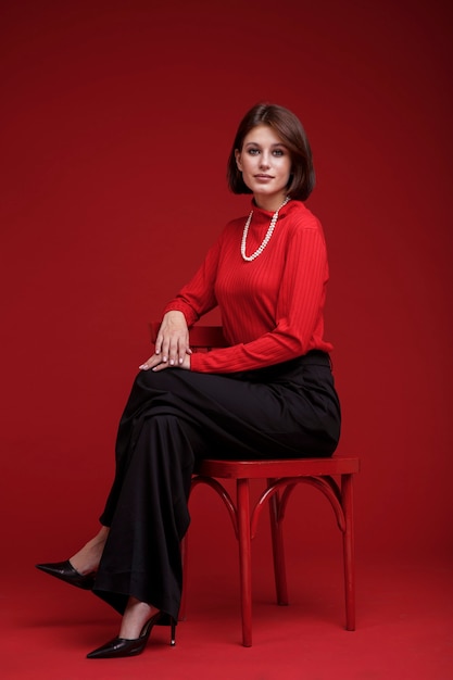 Женщина в черных брюках красный топ жемчужное ожерелье на красном фоне Стрижка Боб Модель сидит на стуле