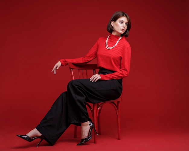 Woman in black pants red top pearl necklace on red background Bob haircut Model sitting on chair