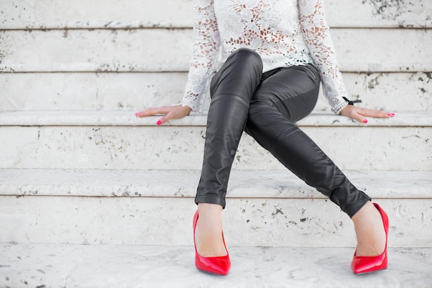 Женщина в черных узких брюках и красных каблуках сидит на лестнице