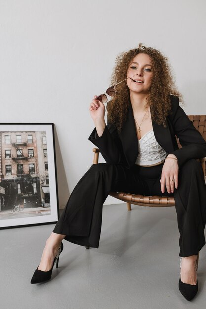 A woman in a black suit sits on a chair and holds glasses