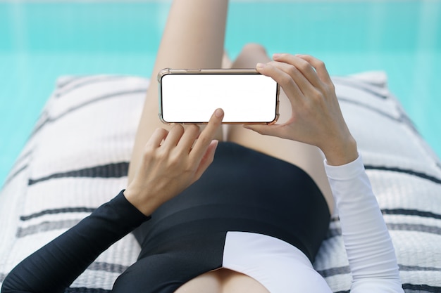 Woman in black and white swimsuit  holding blank mobile phone with white screen near pool.