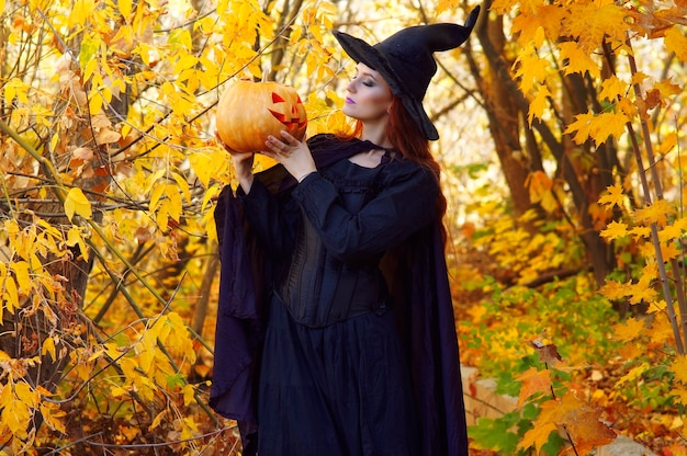 a woman in a black witch costume and a pointed hat with a  pumpkin in a Park on Halloween