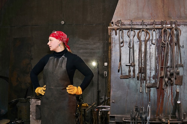 Женщина-кузнец по металлу в своей мастерской рядом с кузнечной печью и инструментами
