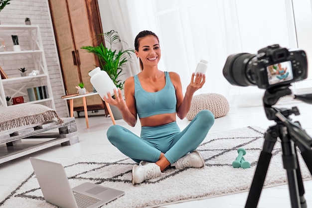 Woman blogger on the floor with dumbbells and a laptop showing a jar of sports nutrition proteins